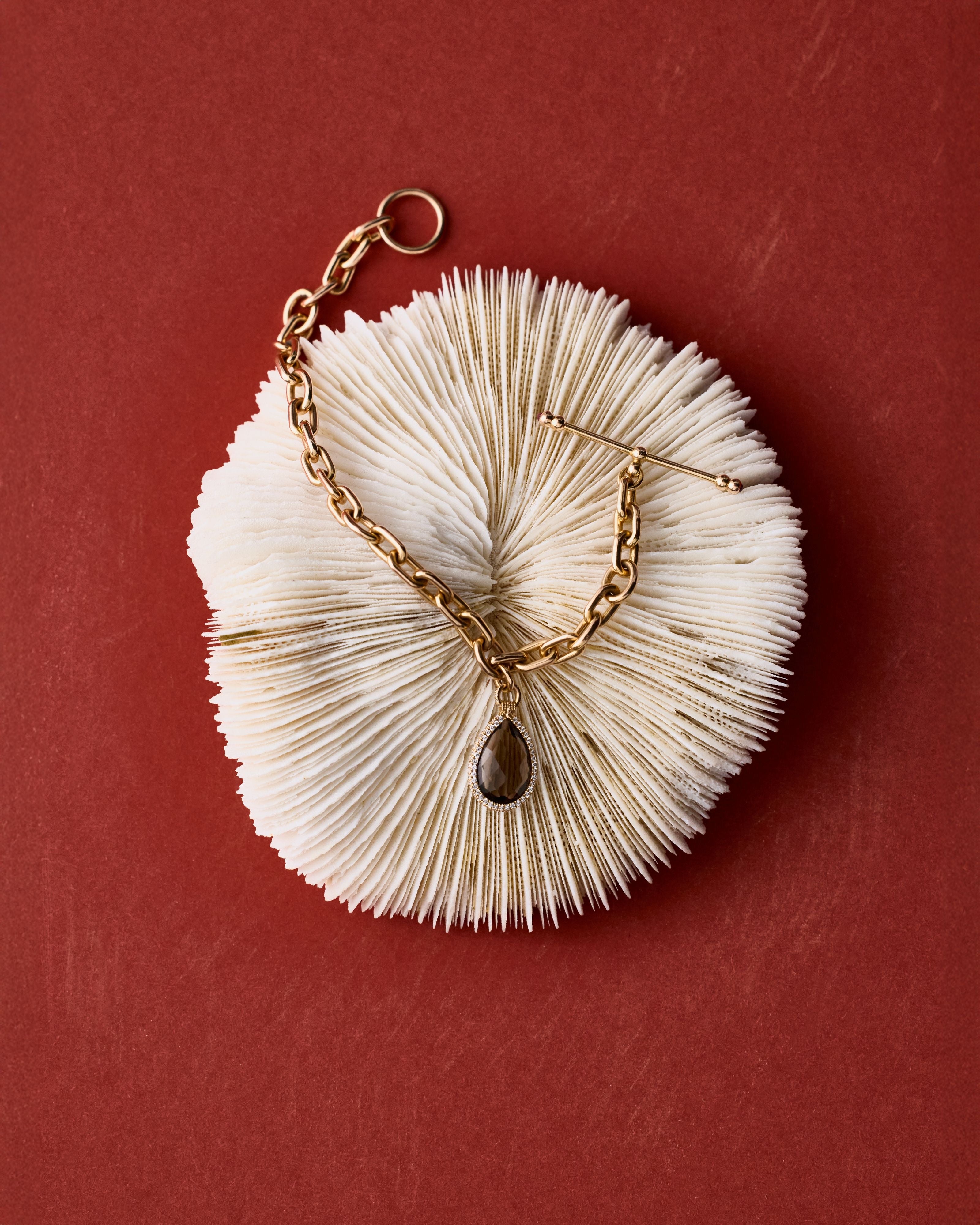 A gold-tone chain necklace with a teardrop-shaped dark brown stone pendant, displayed on a circular, textured white display stand against a solid dark red background.